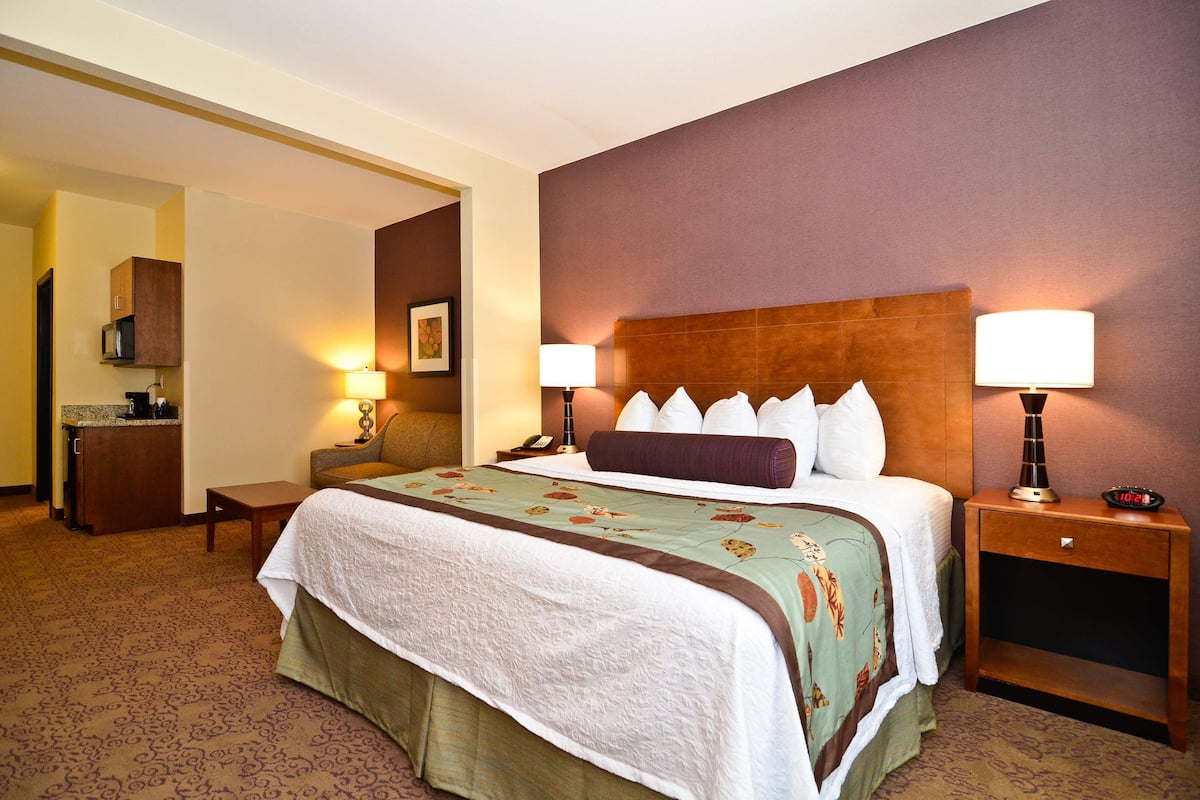 A hotel room with a large bed, two bedside lamps, a nightstand with a clock, a small table, and a microwave in the corner.