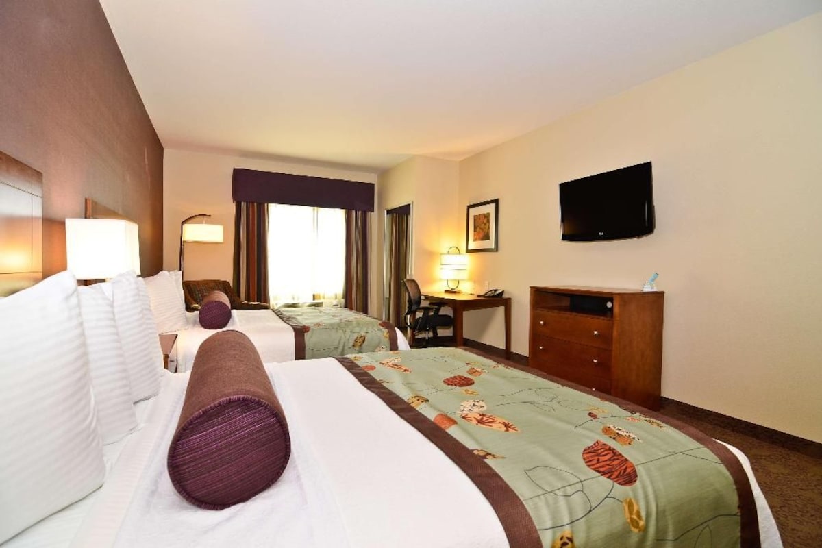 Une chambre d’hôtel avec deux lits, un bureau, une chaise, une télévision et une fenêtre avec des rideaux.
