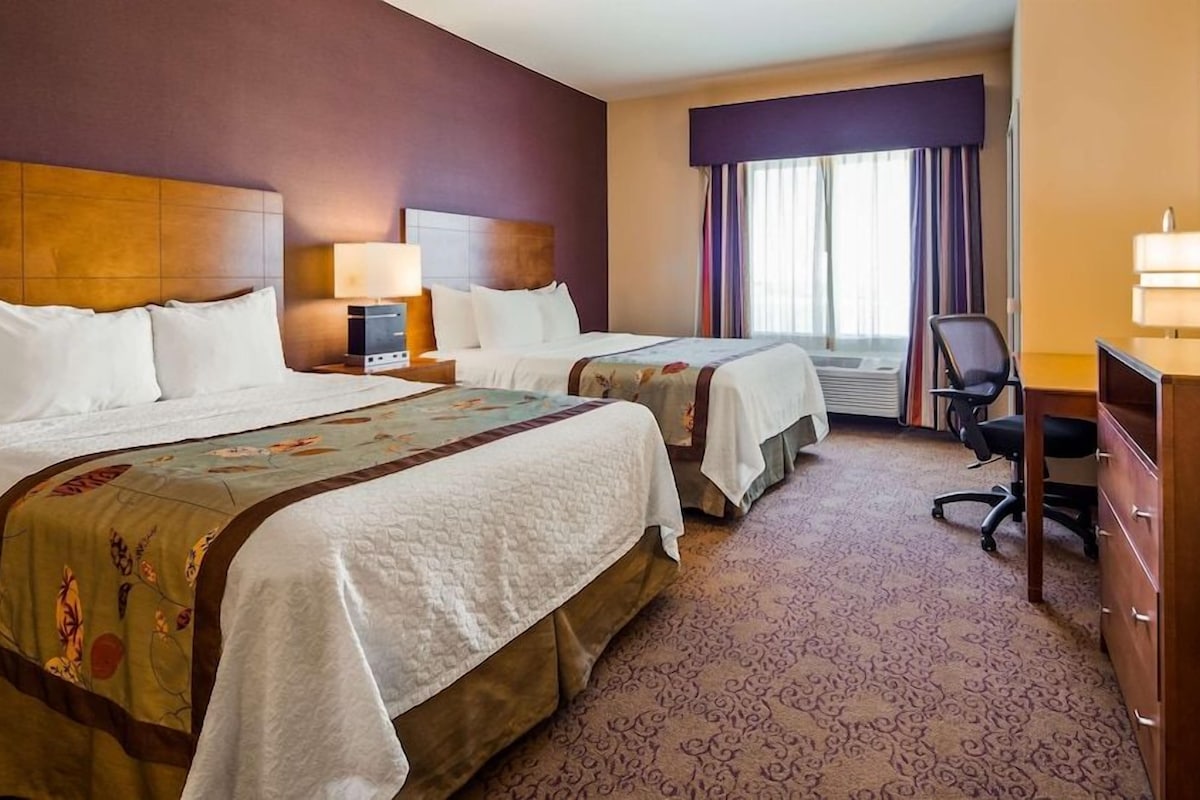 Une chambre d’hôtel avec deux lits, un bureau, une chaise, une commode et une fenêtre avec des rideaux.