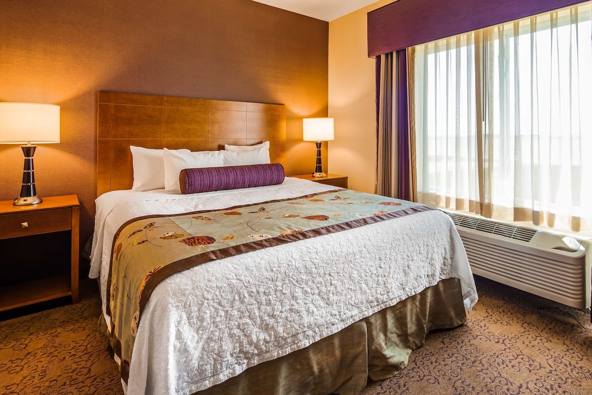 A hotel room with a large bed, two bedside lamps, a window with curtains, and a radiator.