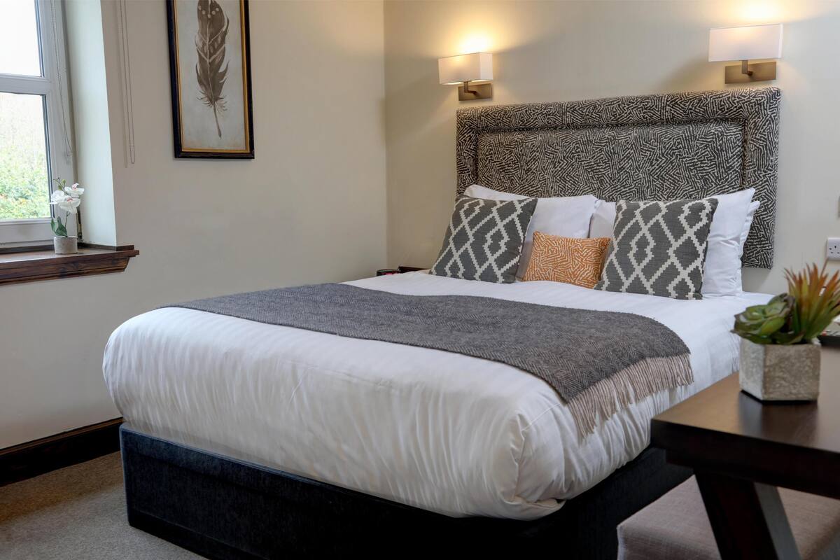 A neatly made bed with a patterned headboard, bedside lamps, and a window with a plant.
