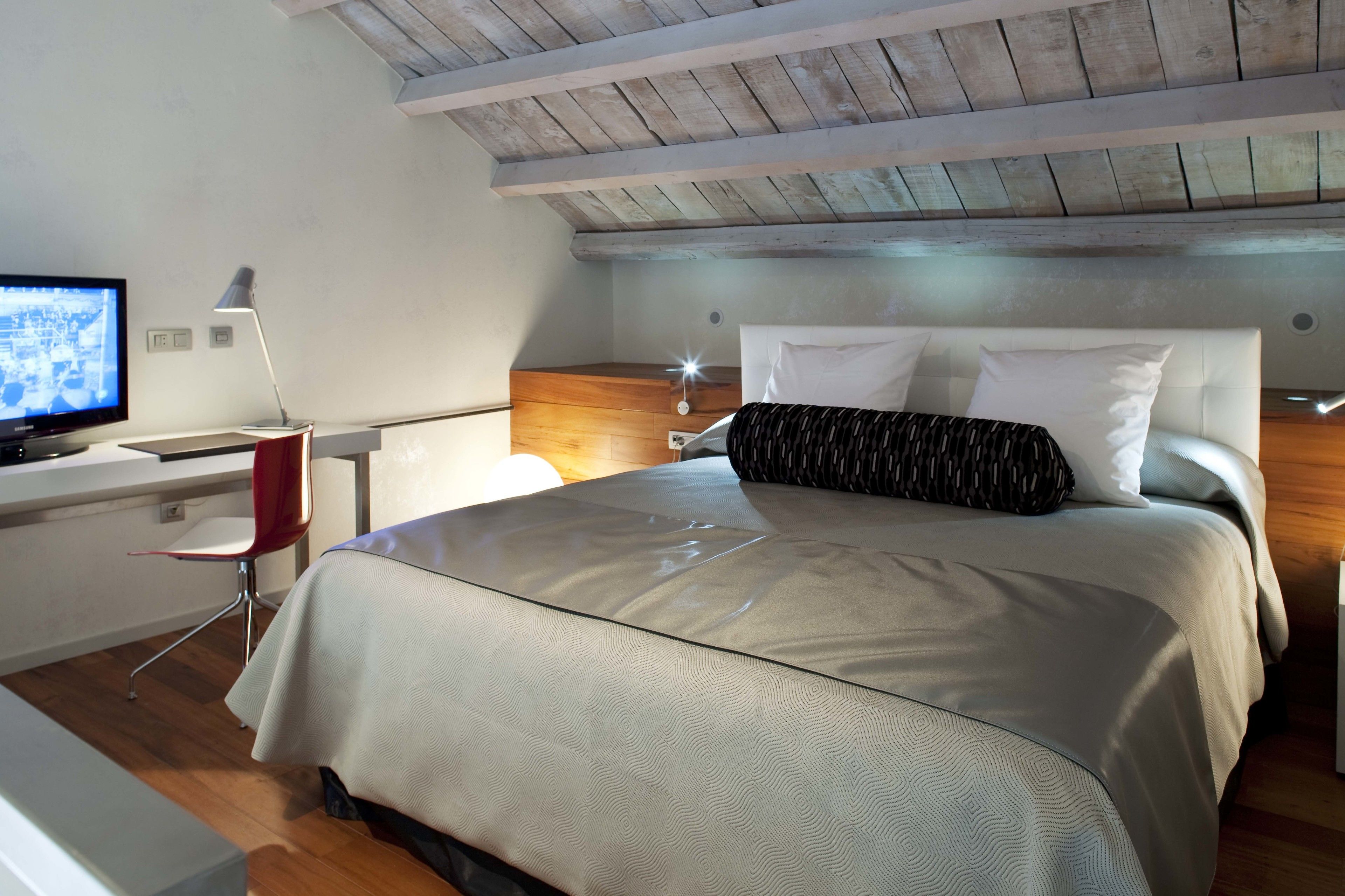 A modern bedroom with a wooden ceiling, a bed with a patterned bedspread, a desk with a computer, and a red chair.