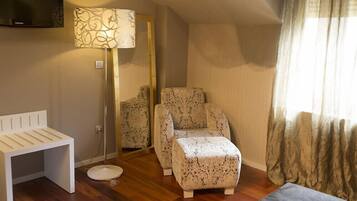 A hotel room with a wooden floor, a white bench, a patterned armchair, a small ottoman, a floor lamp, a mirror, and a bed with a grey bedspread.