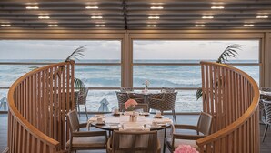 A dining area with a view of the ocean, featuring a round table with chairs, a wooden railing, and a potted plant.