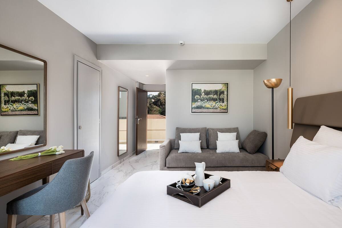 A modern hotel room with a large bed, a desk with a chair, a sofa, and a dining table set with a tray of items.