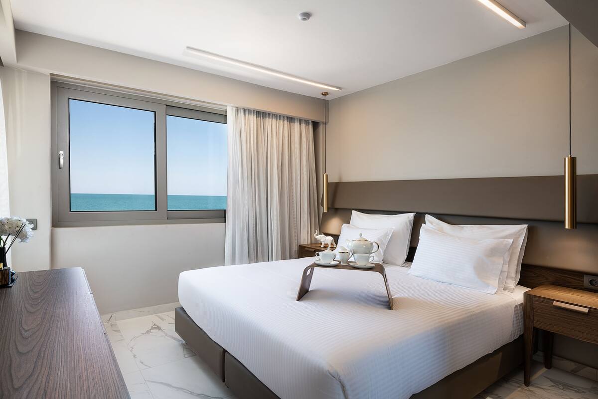 A modern hotel room with a large bed, a bedside table with a lamp, a window with curtains, and a view of the sea.