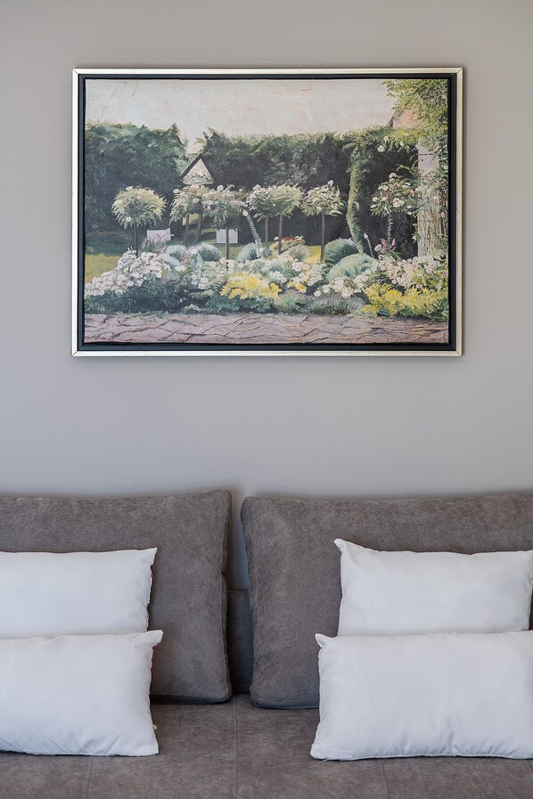 A framed painting of a garden scene on a wall above a grey sofa with white cushions. - Porto Platanias Beach - Luxury Selection (Chania)