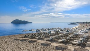 A beach with rows of sun loungers and umbrellas, a clear blue sea, and distant mountains.
