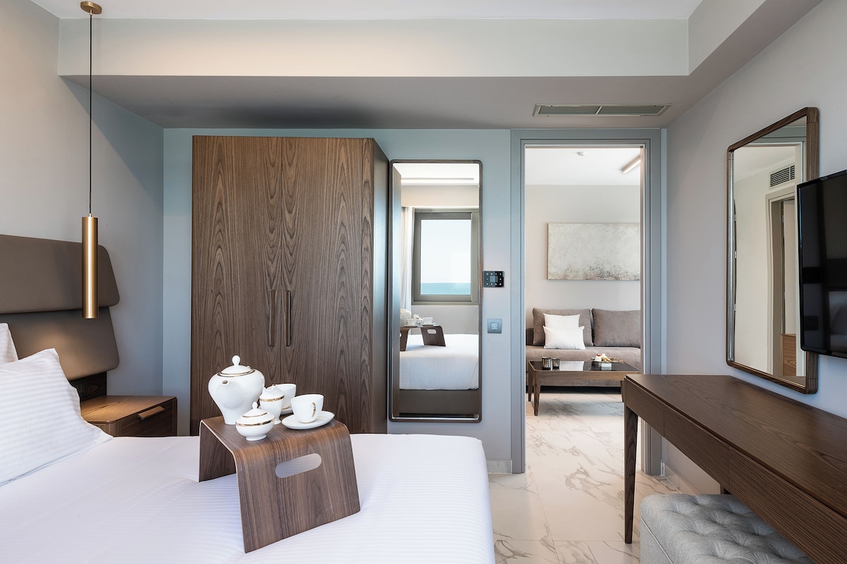 Una habitación de hotel moderna con una cama grande, mesita de noche con una bandeja de teteras blancas, un armario de madera, una zona de estar con sofá y mesa de centro, y un espejo grande.