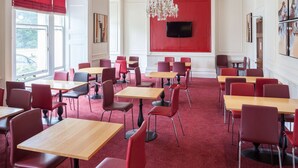 Un espace repas avec des chaises rouges et des tables en bois, un lustre et un mur d’accent rouge orné d’un téléviseur à écran plat.