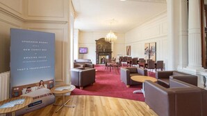 Un hall d’hôtel spacieux, doté d’un tapis rouge, de plusieurs espaces aménagés avec des sièges, d’une cheminée et d’un grand lustre.