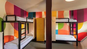 A dormitory room with bunk beds, a wardrobe, and a colorful abstract wall design.