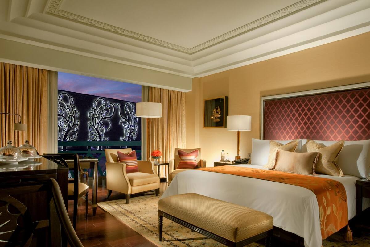 Ein Hotelzimmer mit einem großen Bett, einem Schreibtisch mit Stuhl, einem kleinen Tisch mit Lampe und einem großen Fenster mit Vorhängen.