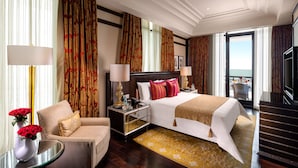 A hotel room with a large bed, two bedside tables with lamps, a chair, a small table with a vase of flowers, and a view of the outdoors. - The Leela Palace Chennai (Chennai)