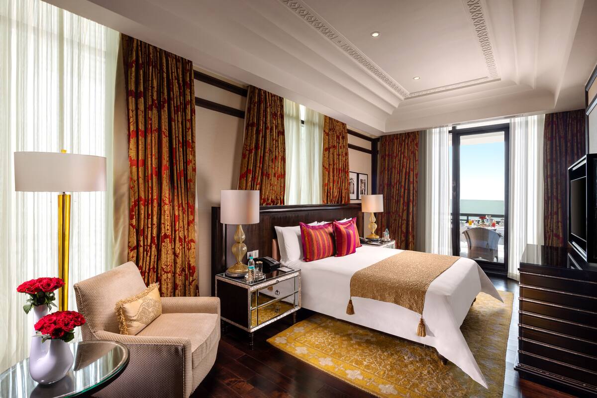 Ein Hotelzimmer mit einem großen Bett, zwei Nachttischen mit Lampen, einem Sessel, einem kleinen Tisch mit Blumenvase und Blick ins Freie.