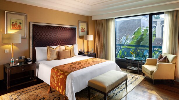 Habitación de hotel con una cama grande, una zona de estar con una silla y una mesa, un balcón con vistas y un techo decorativo.