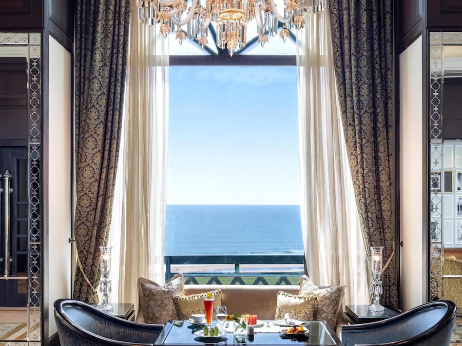 A luxurious room with a large chandelier, a dining table with chairs, and a view of the ocean through large windows.