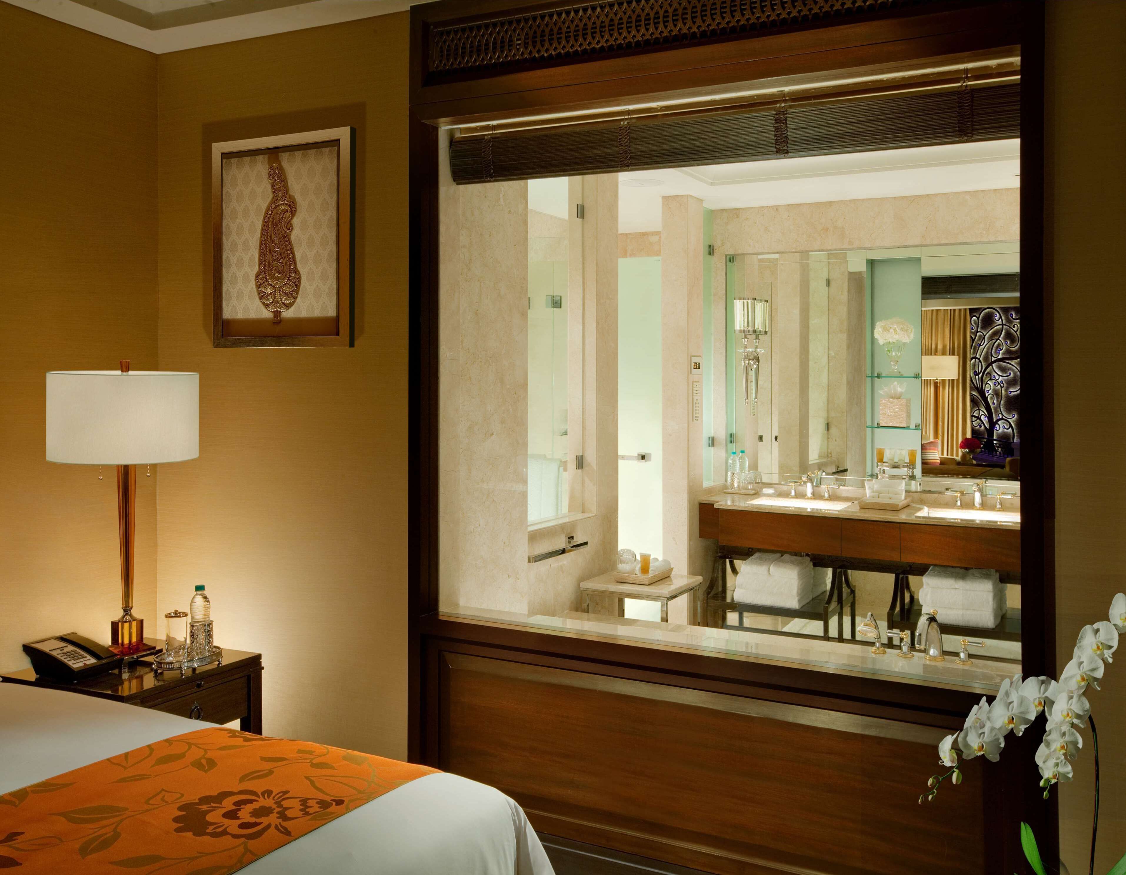 A hotel room with a bed, a bedside table with a lamp, a large mirror reflecting the bathroom, and a floral decoration.
