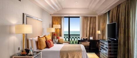 A spacious hotel room with a large bed, two bedside tables, a desk, and a television. The room features a balcony with a view of the ocean.