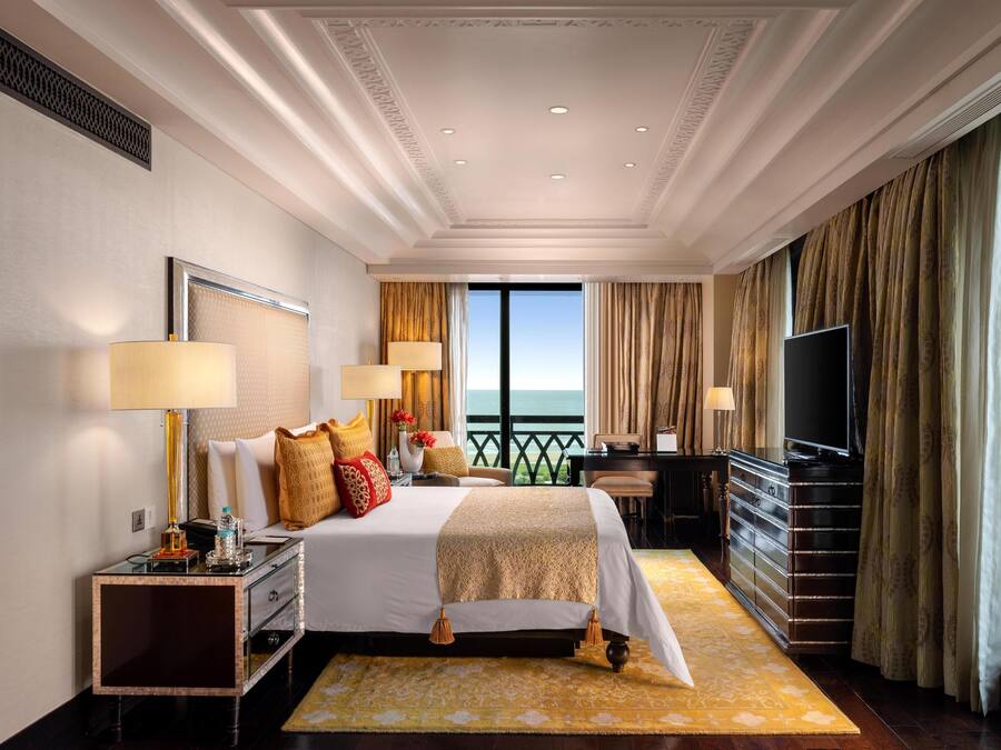 A spacious hotel room with a large bed, two bedside tables, a desk, and a television. The room features a balcony with a view of the ocean.