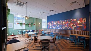 A restaurant with wooden flooring, multiple seating areas including tables and booths, hanging pendant lights, and a large abstract mural on the back wall. - Travelodge Brighton Seafront (Brighton)