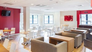 A modern dining area with wooden tables and white chairs, a red accent wall, and a large window providing natural light. - Travelodge Maidstone Central (Maidstone)
