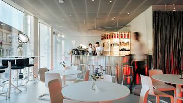 A modern bar area with white tables and chairs, a bar counter with bottles, and a large window with natural light.