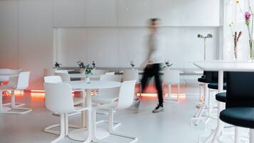 A modern, well-lit conference room with white tables and chairs, a person walking by, and a vase with flowers on a table.