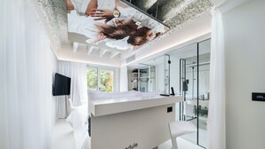 Habitación de hotel moderna con escritorio blanco, televisión de pantalla plana y ventana grande con vistas a zonas verdes.