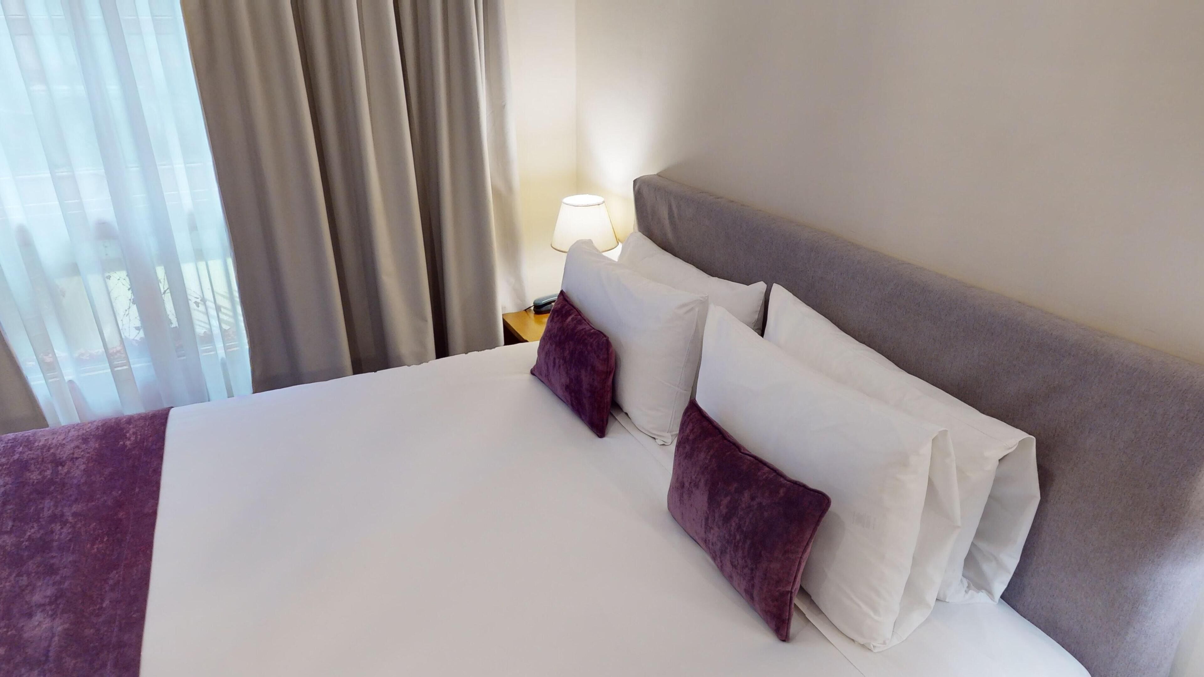 A neatly made bed with white and purple bedding, a grey headboard, and a bedside lamp.