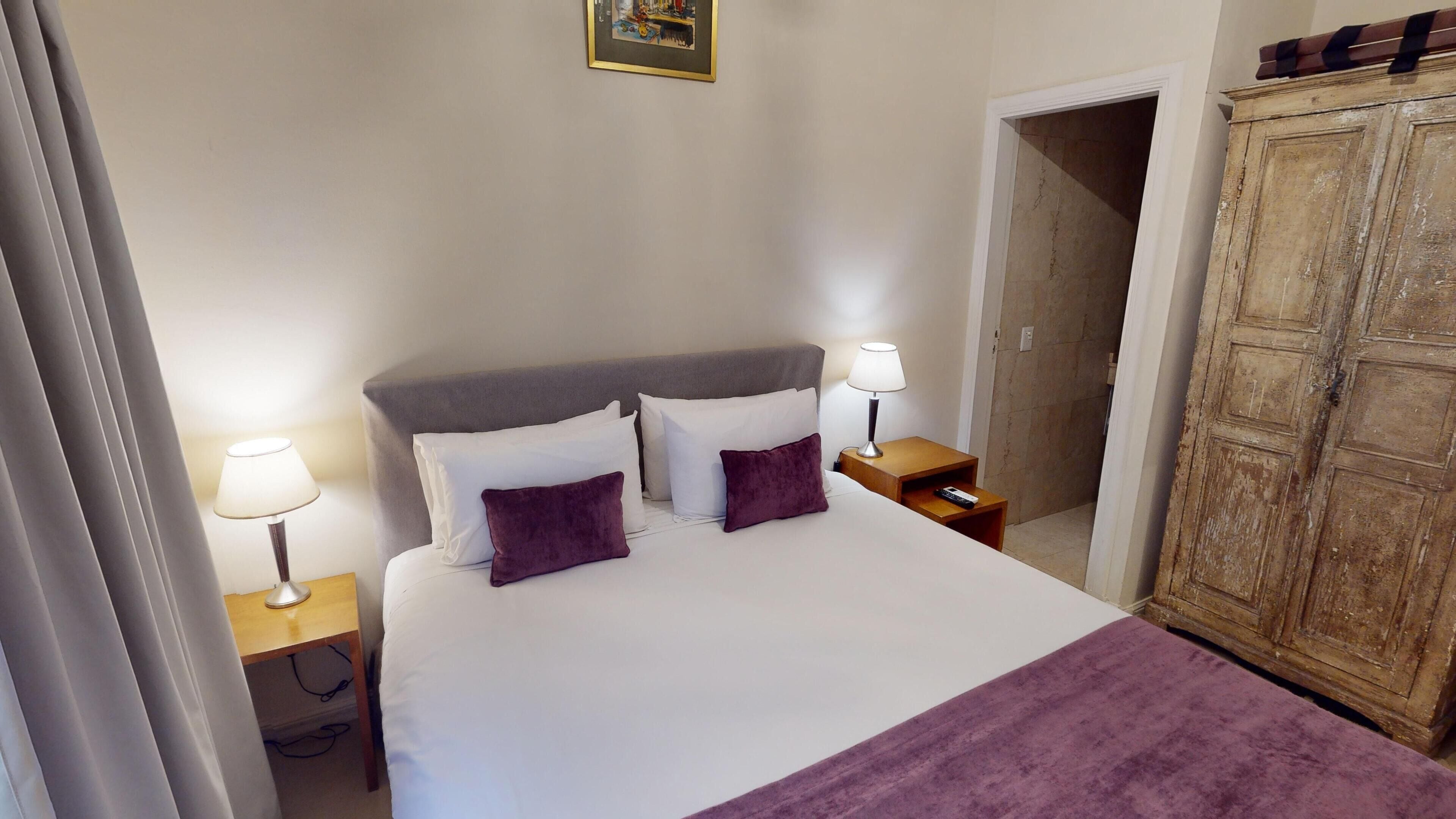 A neatly made bed with white linens and two purple pillows, flanked by bedside tables with lamps. A wooden wardrobe is visible to the right.