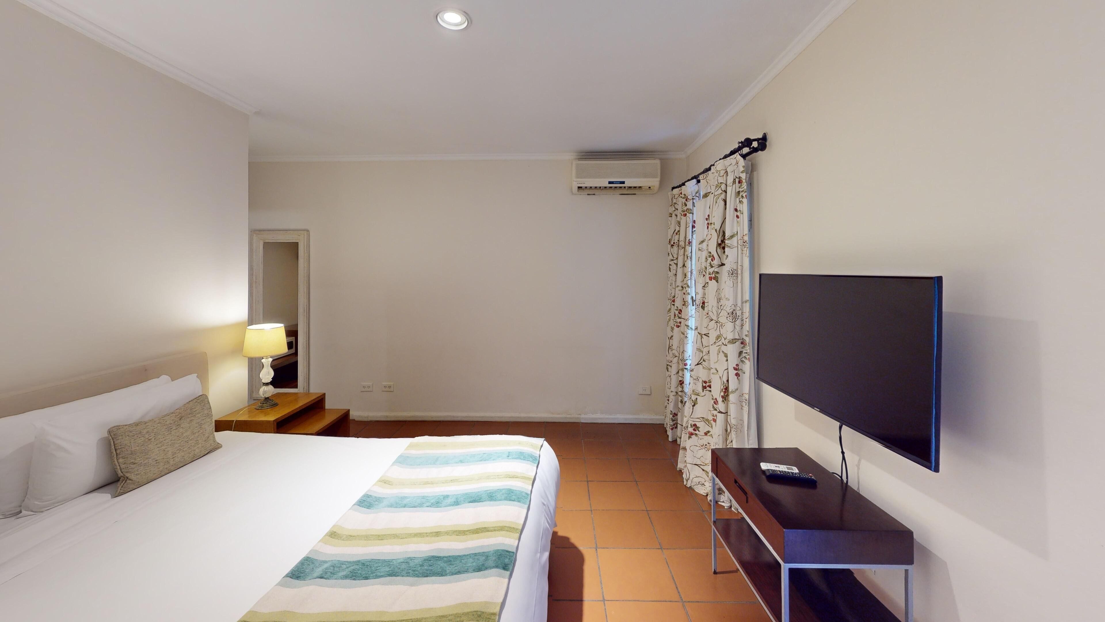 A hotel room with a bed, a bedside table with a lamp, a TV on a stand, and an air conditioning unit on the wall.