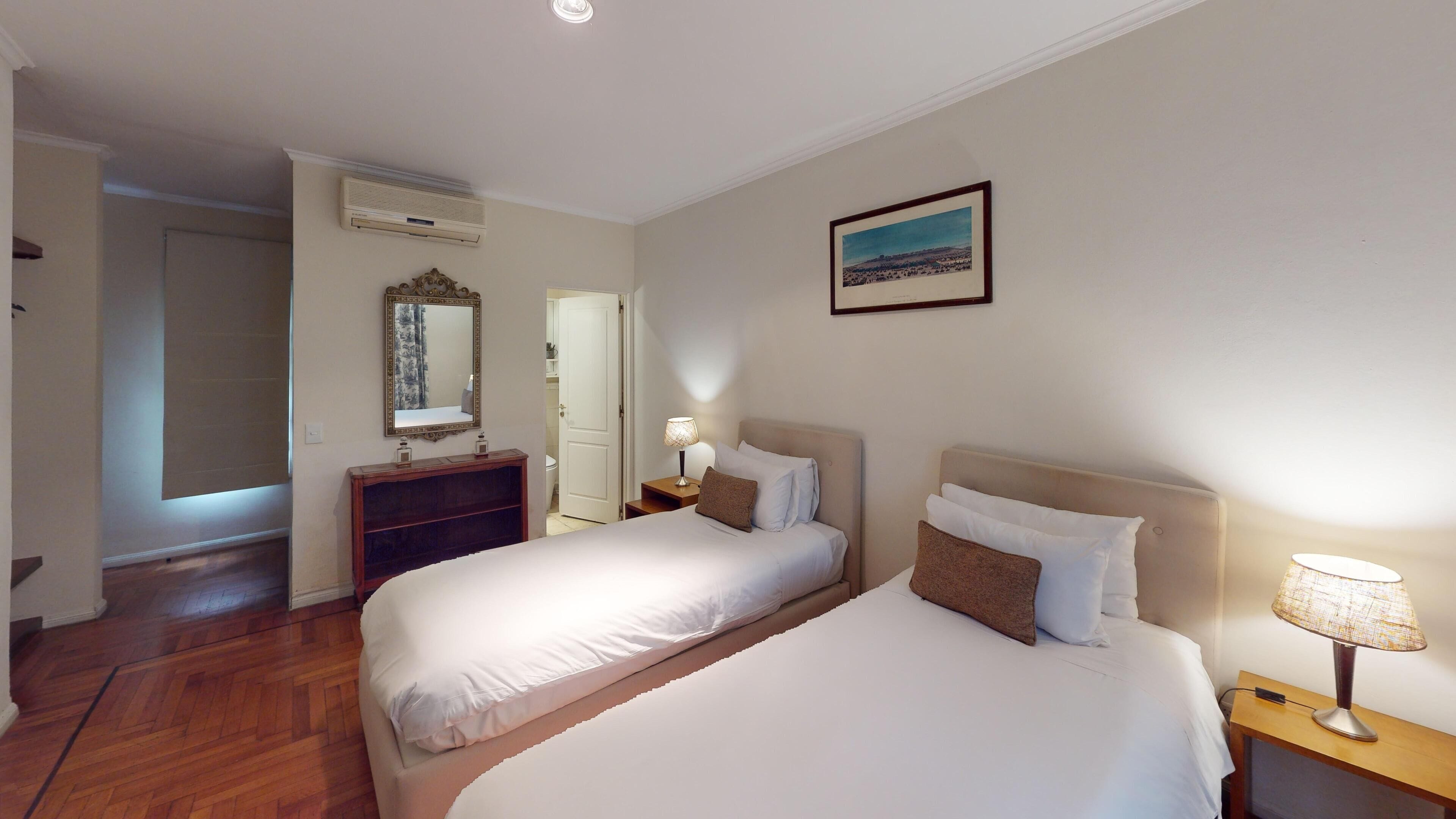 Una camera d'albergo con due letti, un comò in legno, uno specchio, un comodino con una lampada e un quadro appeso al muro.