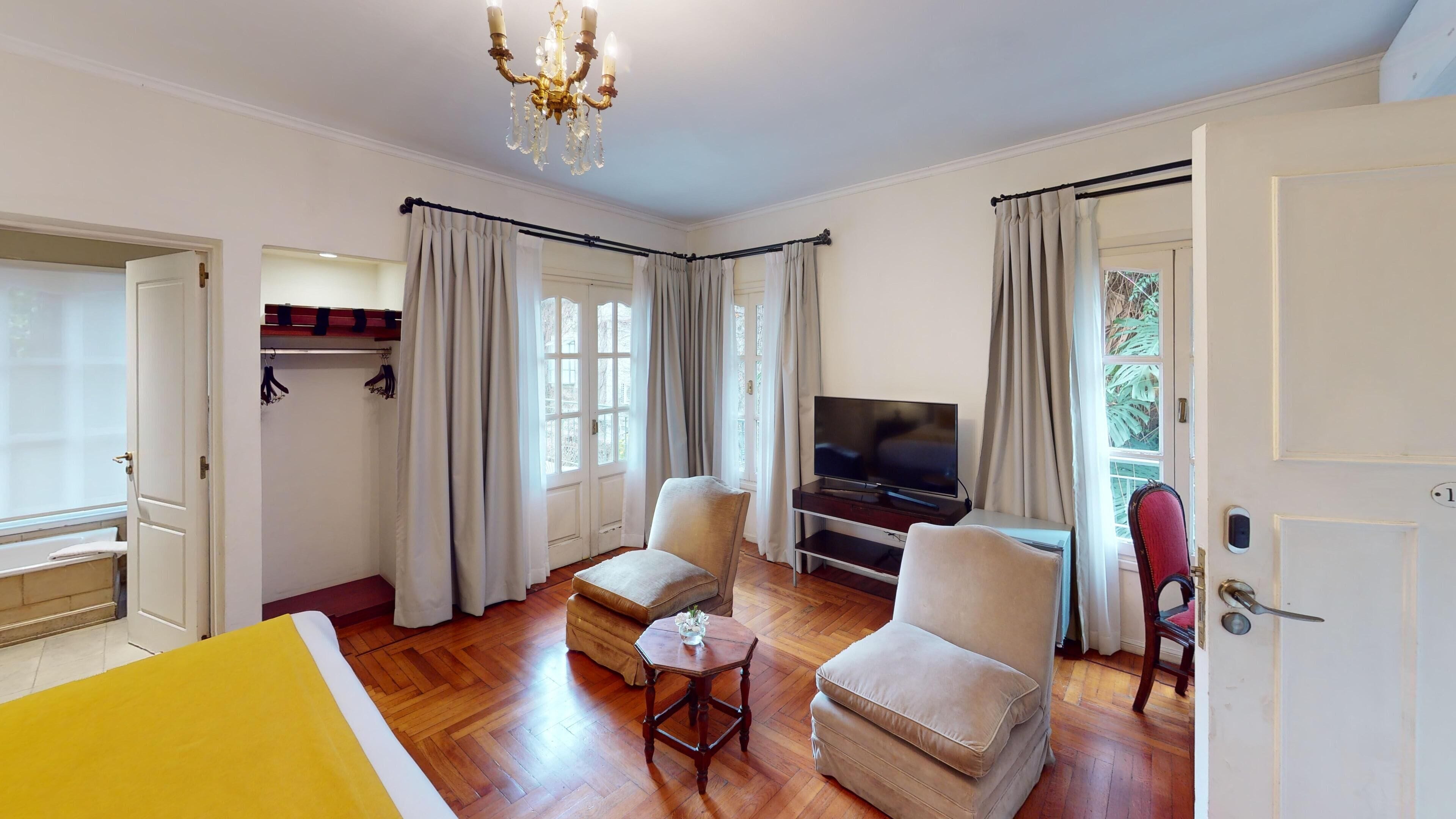 A spacious room with a bed, two armchairs, a small table, a TV, and a chandelier.