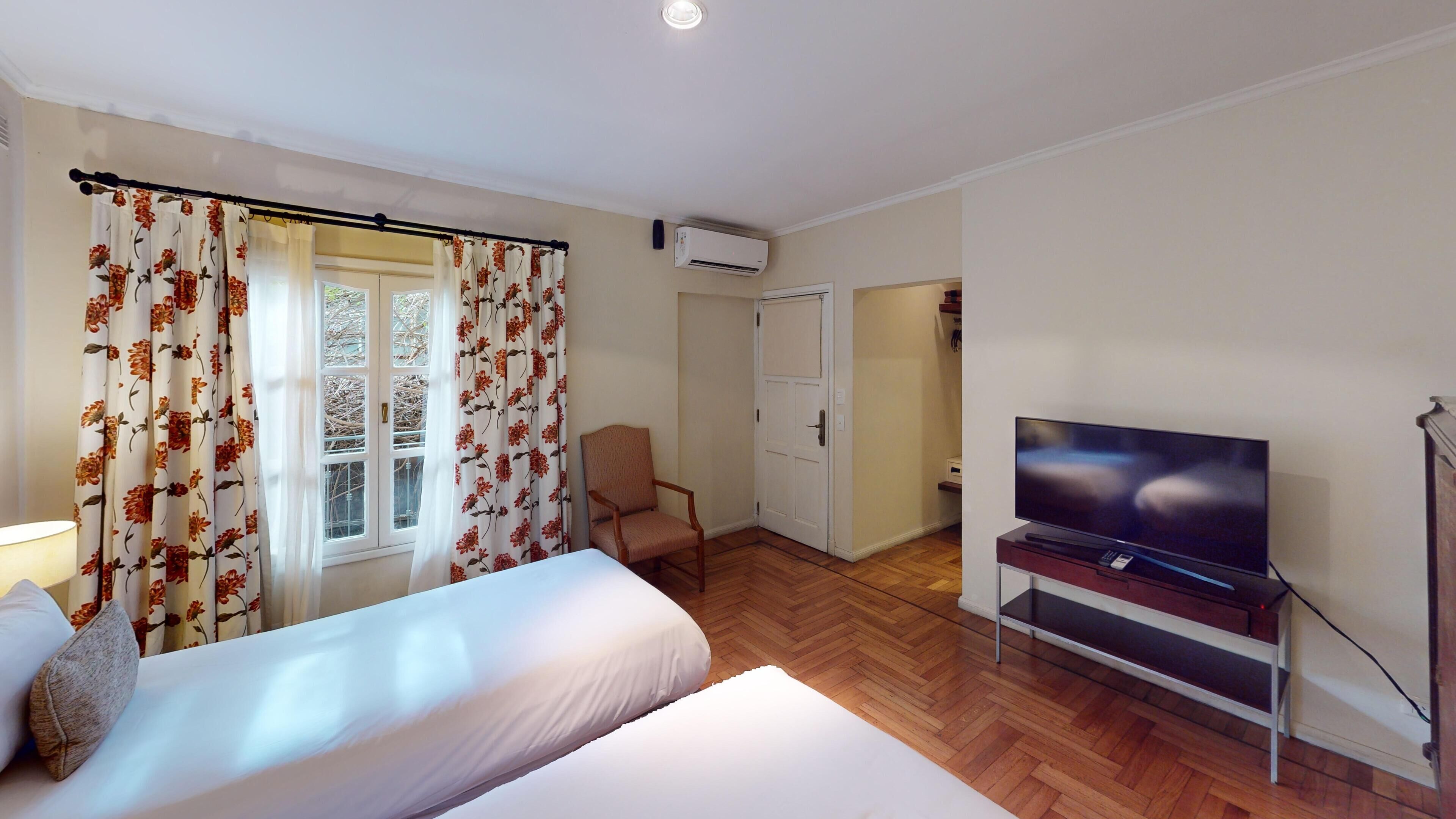 Quarto de hotel com cama, televisão, cadeira e janela com cortinas florais.