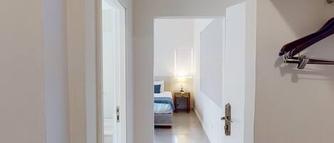 A narrow hallway leading to a bedroom with a bed, bedside table, and a lamp.