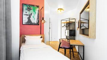 Une chambre d’hôtel moderne, équipée d’un lit, d’un bureau et d’une chaise. Un tableau est accroché au mur et un miroir se trouve au-dessus du bureau.
