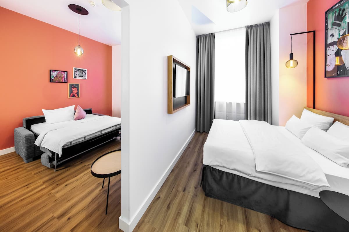 A modern hotel room with a wooden floor, two beds, a small table, and a large window with curtains.