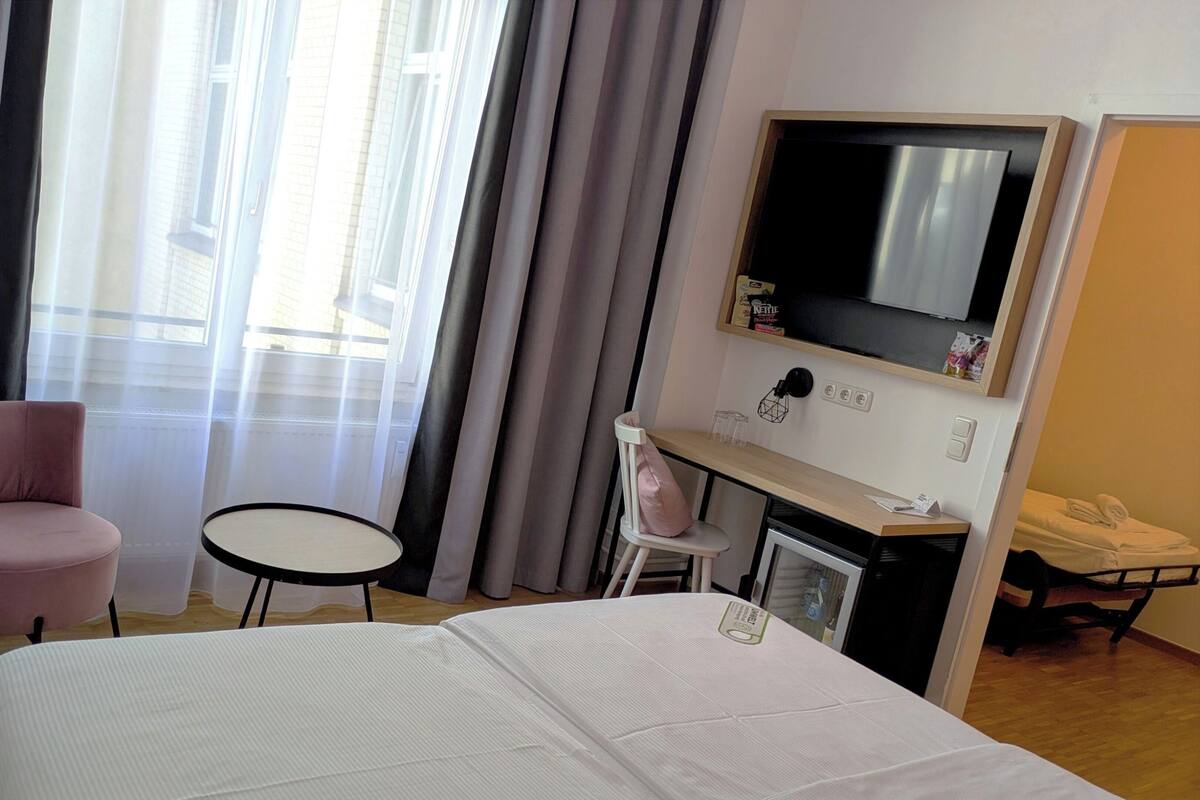 A hotel room with a bed, a pink chair, a small table, a TV, and a desk with a kettle and other items.