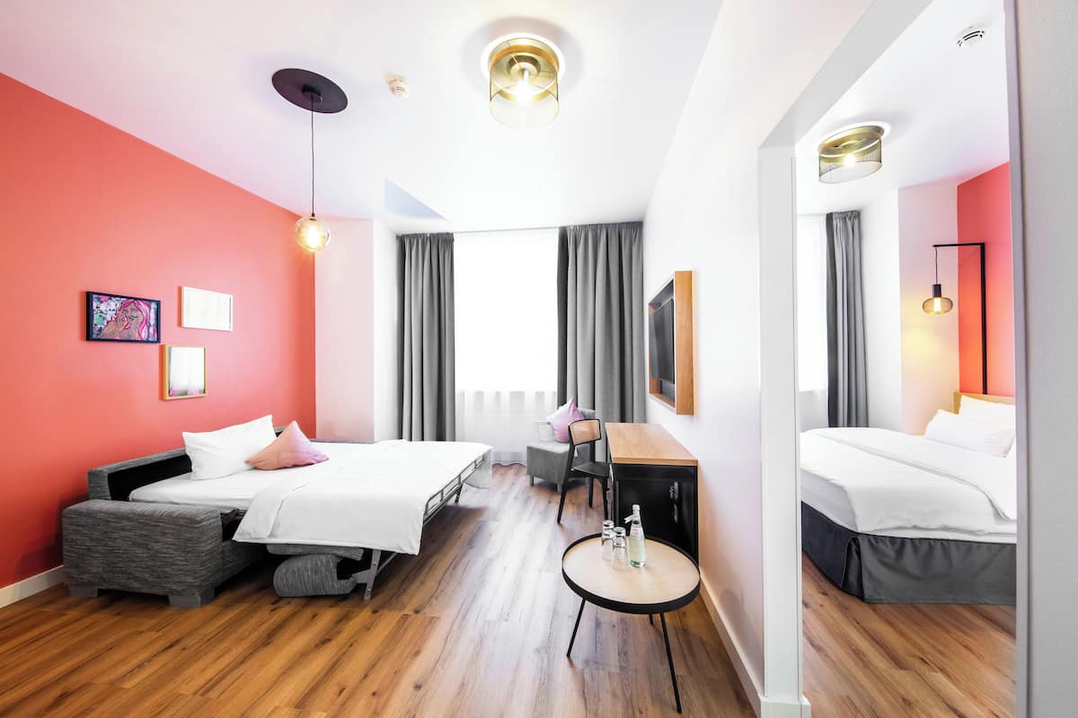 A modern hotel room with a wooden floor, a bed with a grey upholstered headboard, a desk with a chair, and a small round table with a bottle and glasses.