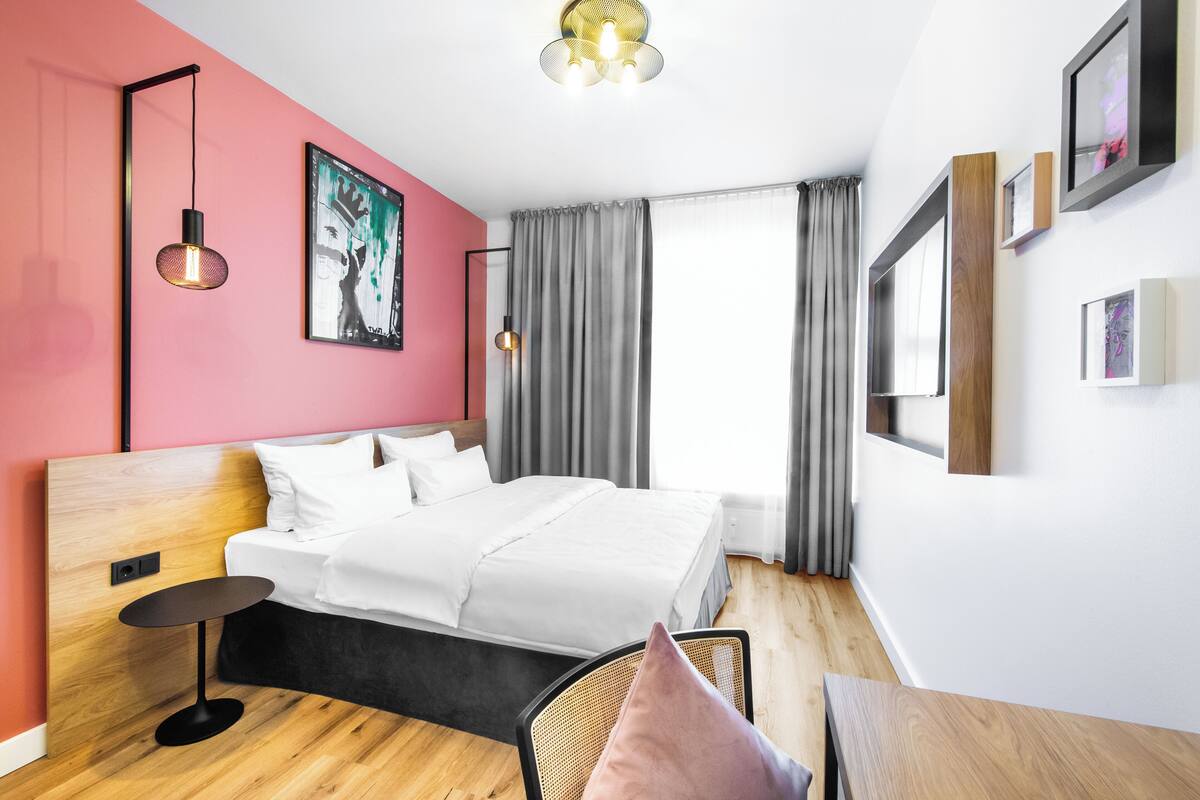 A modern hotel room with a wooden bed, a small side table, a chair, and a large window with curtains.