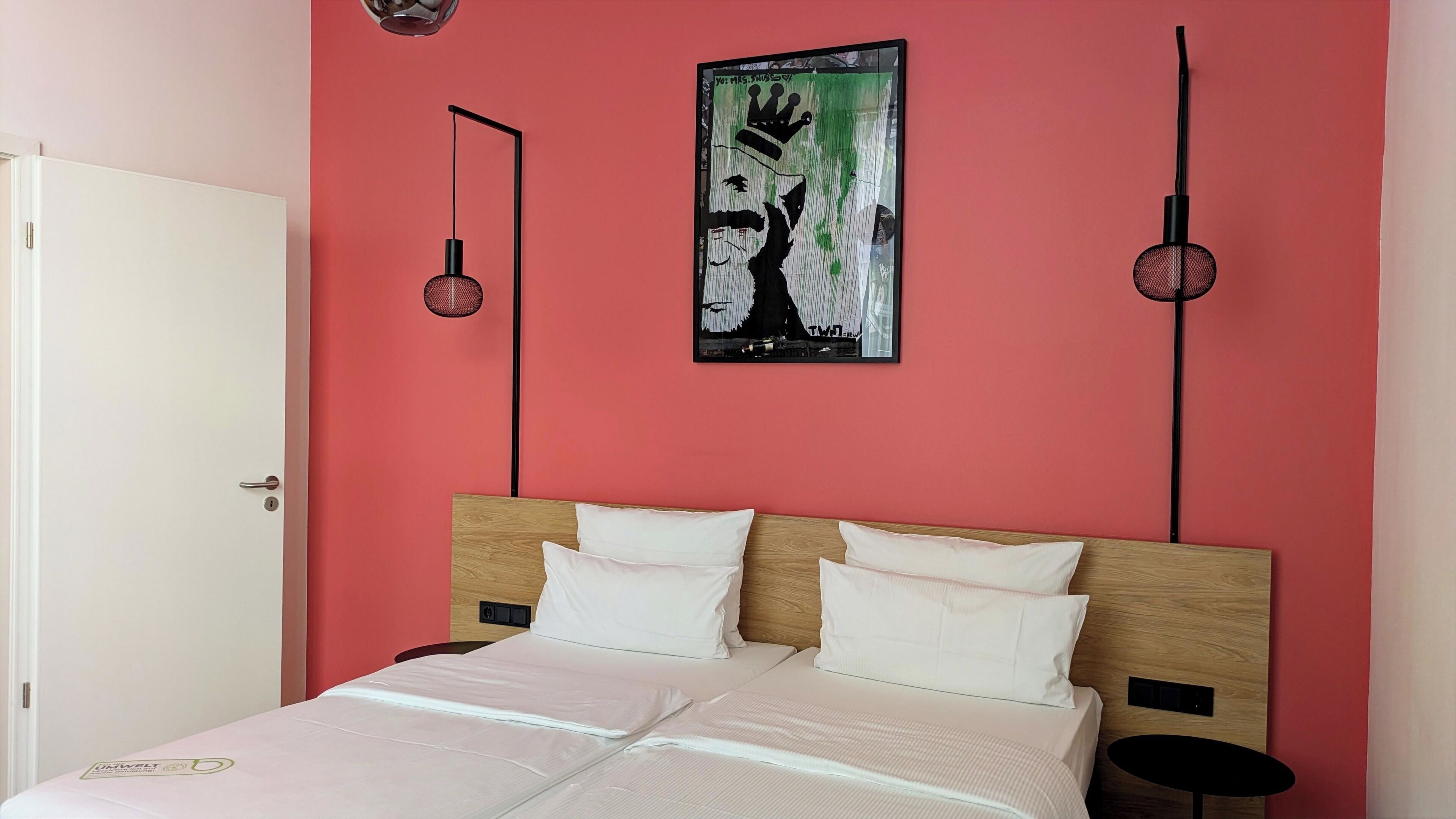 Ein Schlafzimmer mit roter Wand, einem Bett mit weißer Bettwäsche, einem hölzernen Kopfteil, einer schwarzen Stehlampe und einer kleinen schwarzen Tischlampe, sowie einem gerahmten Kunstwerk über dem Bett.
