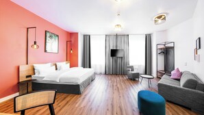 A modern hotel room with a bed, a sofa, a chair, a small table, and a TV. - CALMA Berlin Mitte (Berlin)