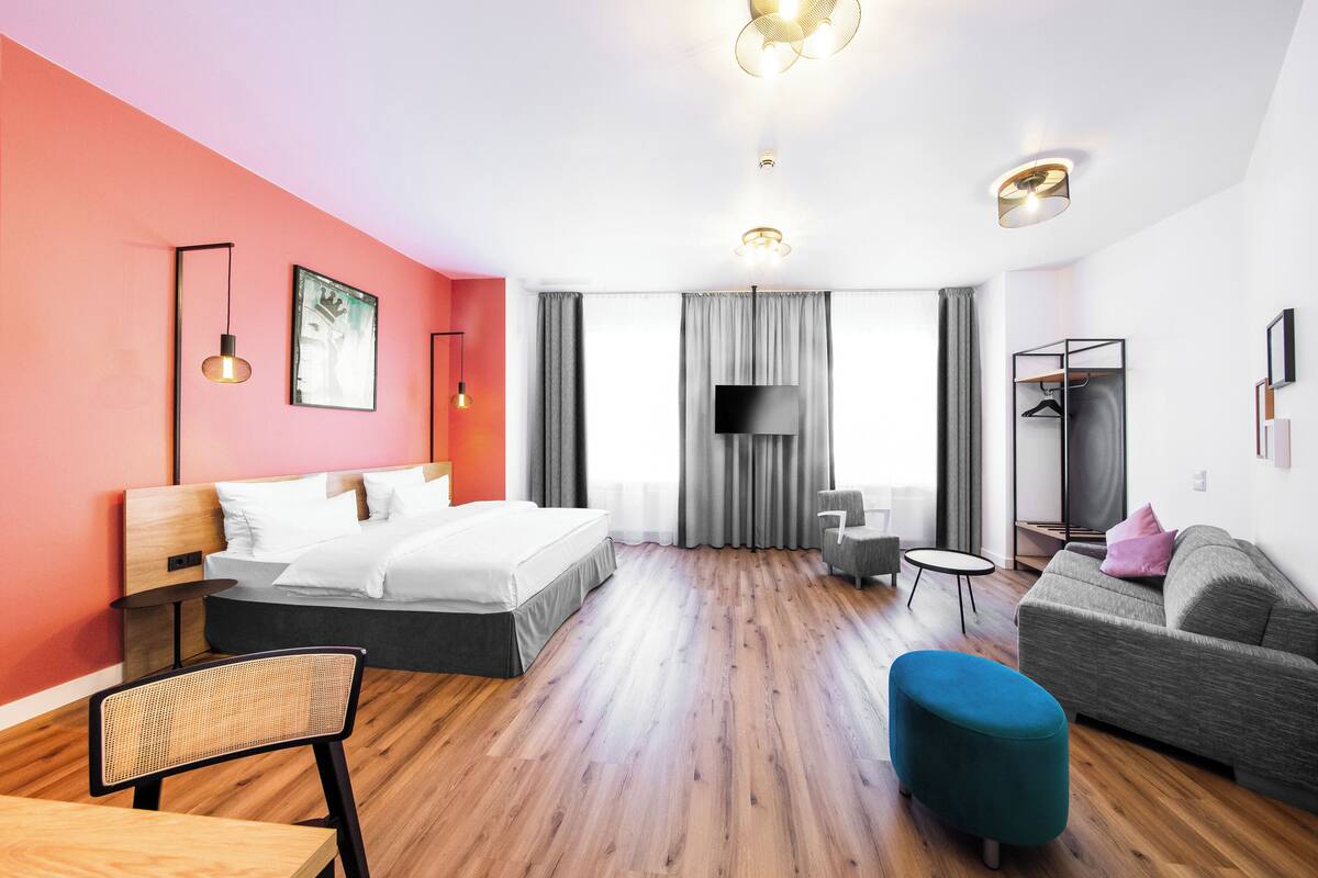 A modern hotel room with a bed, a sofa, a chair, a small table, and a TV.