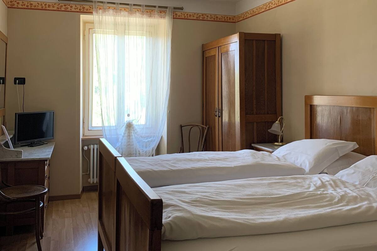 A bedroom with two beds, a desk with a computer, a chair, a wardrobe, and a window with sheer curtains.