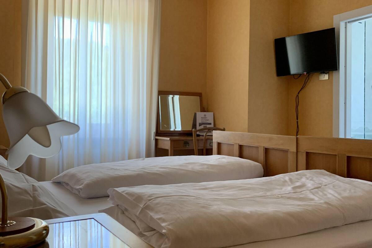 A hotel room with two beds, a desk, a lamp, a television on the wall, and a window with curtains.