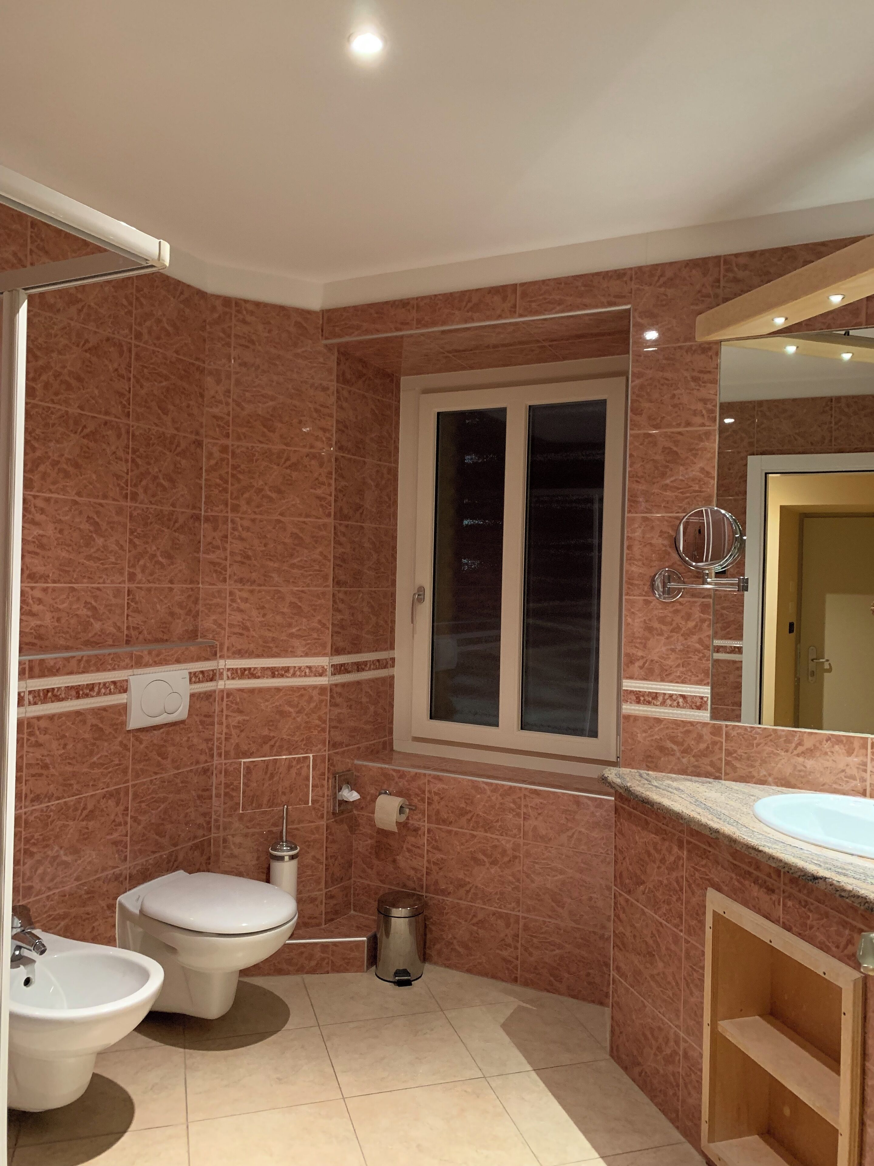 A bathroom with a toilet, bidet, and sink, marble walls, and a window with a mirror.