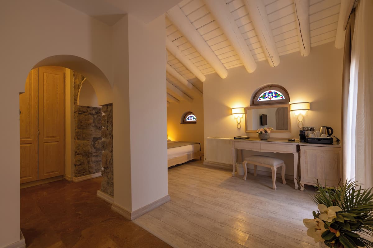 Una habitación con suelo de madera, pared de piedra, puerta de madera, una banca, un escritorio, un espejo y una ventana con diseño de vitral.