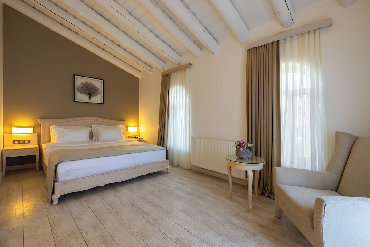Un dormitorio con cama, lámparas de noche, una silla, una mesita y una ventana con cortinas.