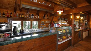 A rustic bar area with wooden beams, a variety of bottles, and a display case with fruits and pastries. - Chalet del Sole (Sauze d'Oulx)